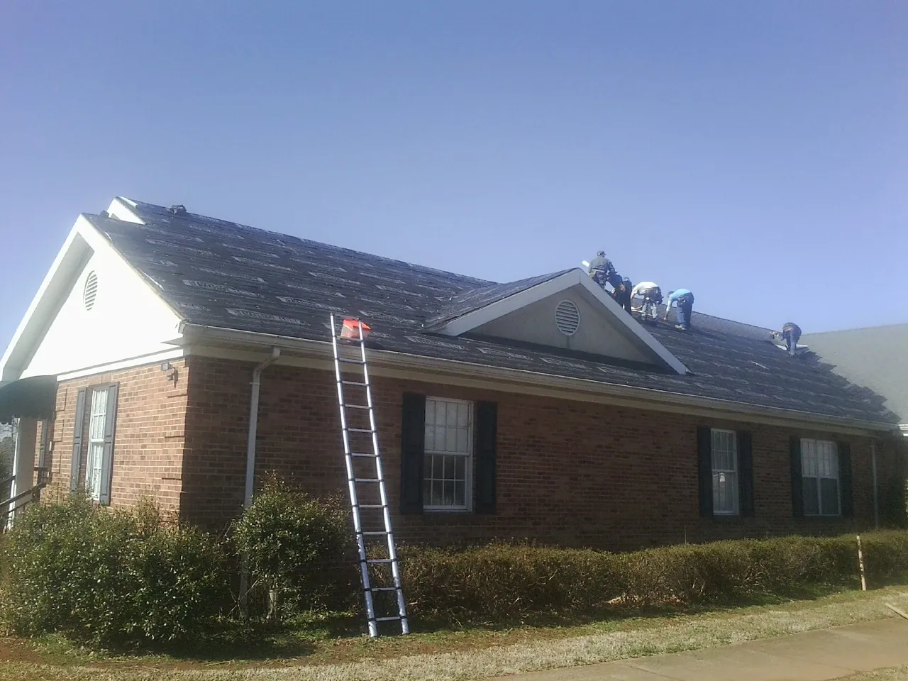 Crew tearing shingles off a brick ranch roof with a ladder set — Metal Roof Repair work by our crew in Pond Eddy