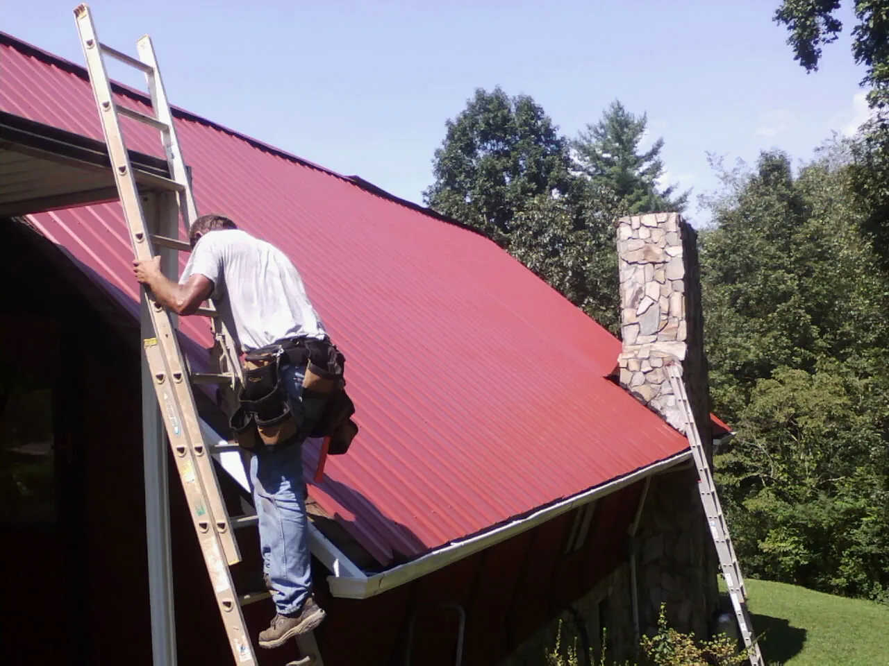 Roofer on a ladder beside a red metal roof and stone chimney — Soffit & Fascia Repair by Roofing Brothers in Pond Eddy
